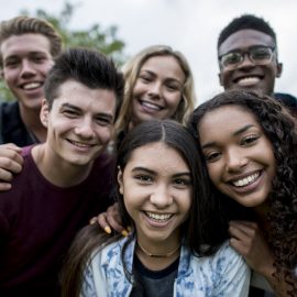 A multi-ethnic group of teenagers are outdoors on a cloudy day. They are wearing casual clothing. They are smiling while taking a selfie together.